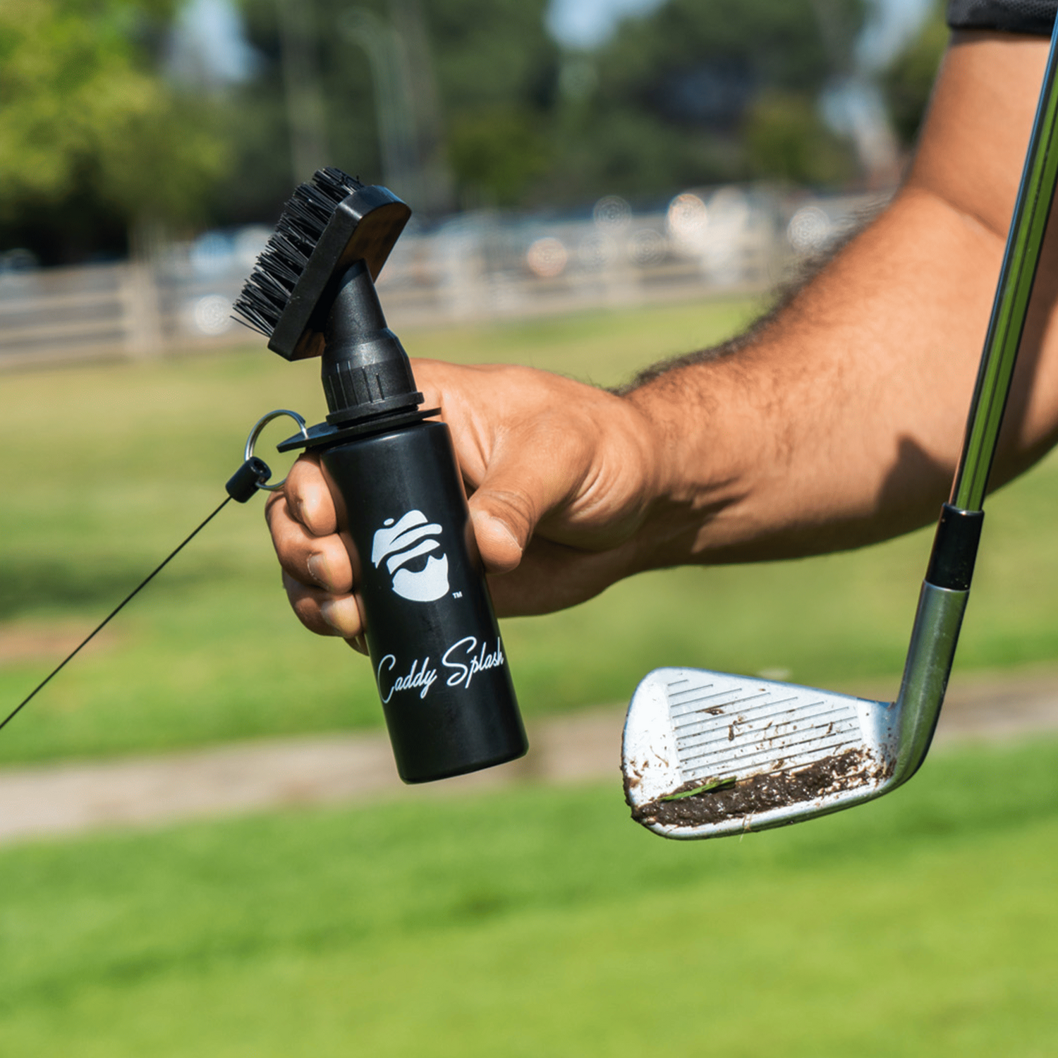 Person holding a golf club and a black bottle with a brush attachment on a golf course.